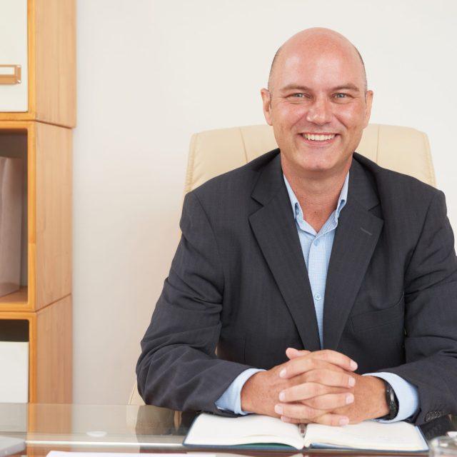 Portrait of mature smiling businessman in black jacket sitting at his workplace at office