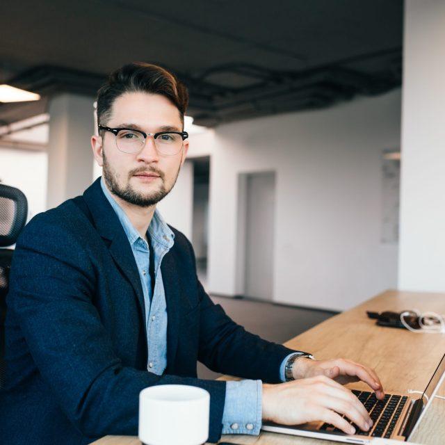 Young dark-haired man is working at the table in office. He wears blue shirt with black jacket. He is typing on laptop and looking to the camera.